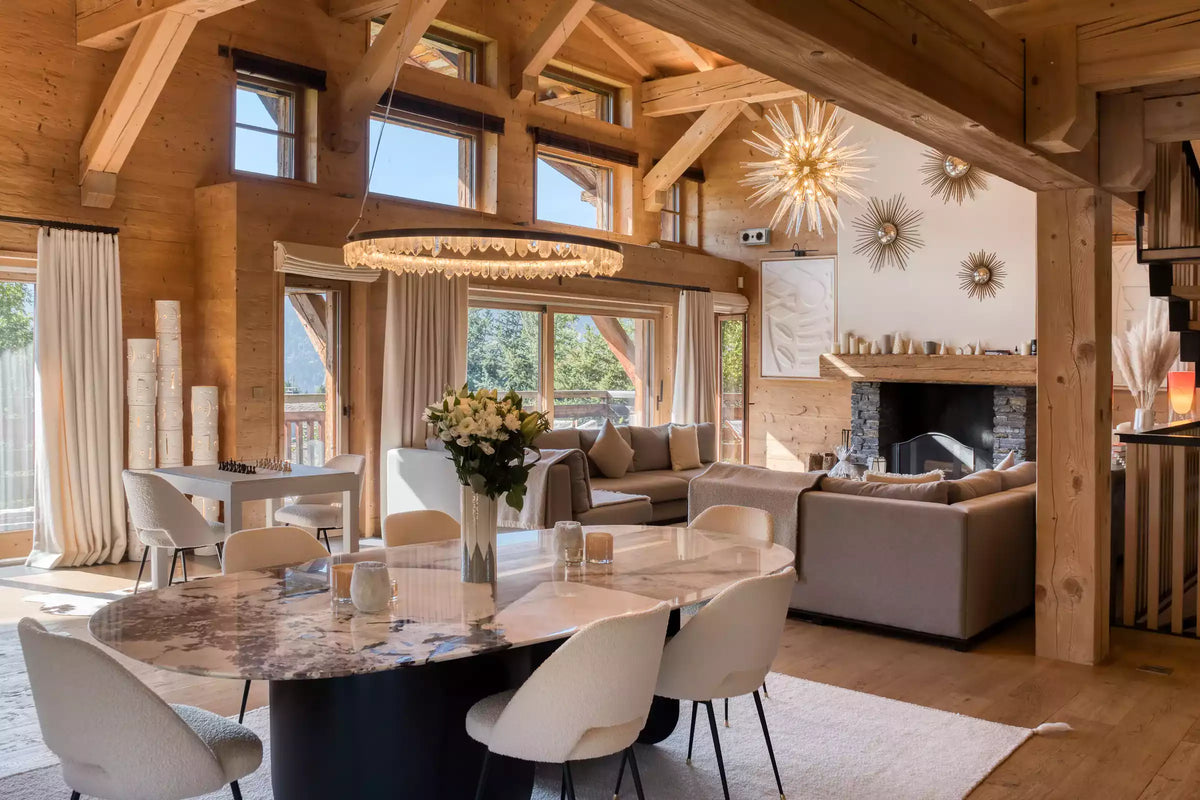 A luxury chalet-style dining and living room with high wooden beams, a marble-top dining table with white chairs, a fireplace, and large windows looking out onto a mountain view.
