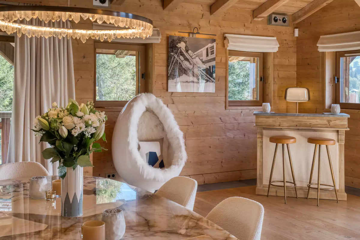 A modern interior room with wood-paneled walls, a large white egg-shaped chair with fluffy trim, a marble dining table with chairs, a floral centerpiece, and a small bar area with two wooden stools.