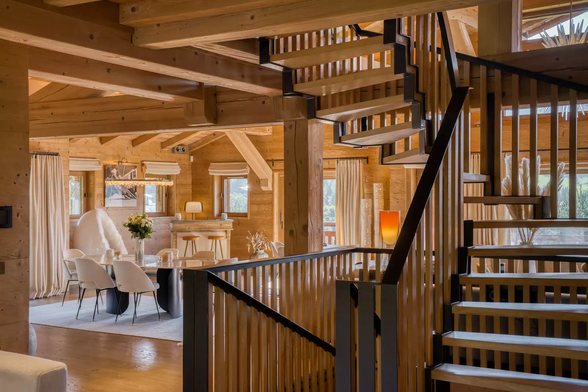 A modern wooden staircase with black metal railings in a rustic log cabin interior, overlooking a dining area with light-colored chairs and warm wood walls.