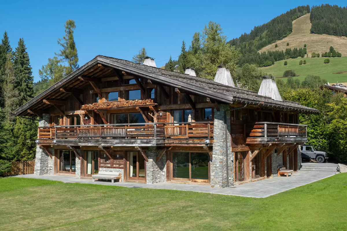 A large alpine-style wooden chalet with stone accents, multiple balconies, a sloped dark roof, and a paved courtyard, set on a grassy lawn with trees and a mountain in the background.