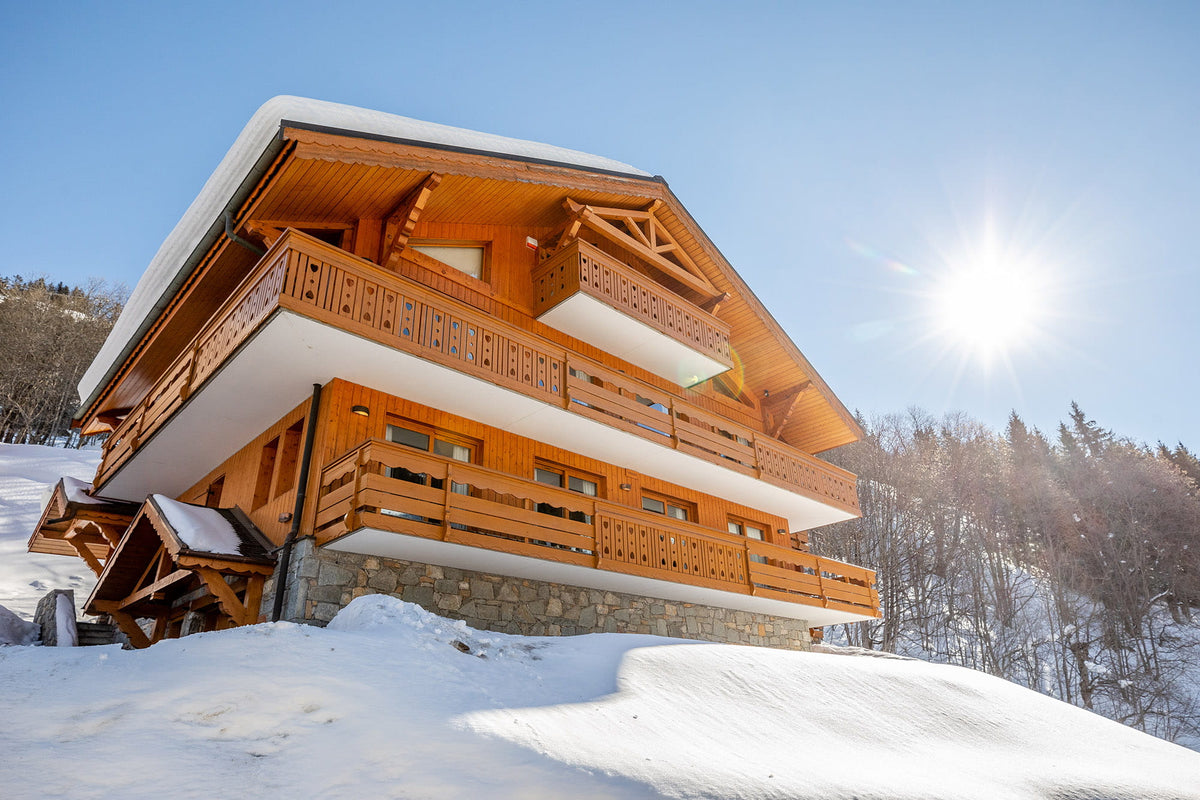 A traditional wooden chalet mountain house with snow-covered roofs and stone foundations, set in a snowy landscape under a bright blue sky with the sun shining.