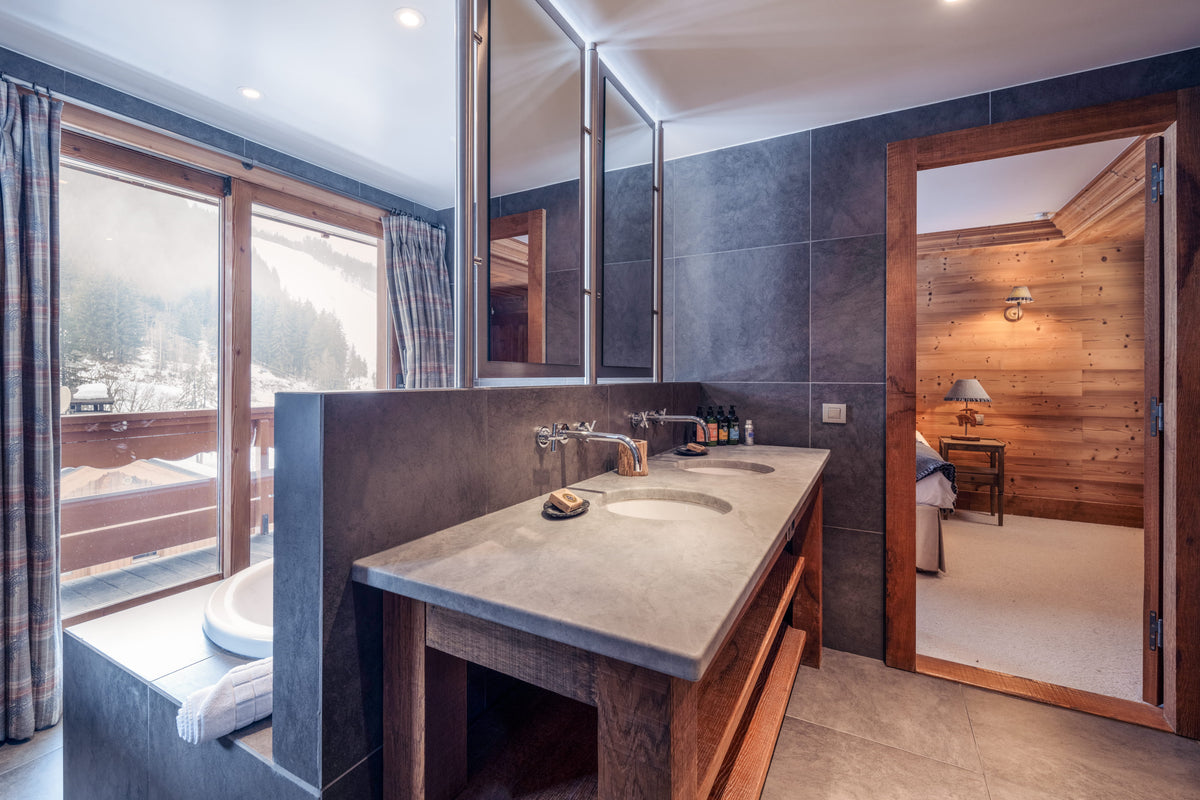 A luxury modern bathroom featuring a double-sink vanity with a stone countertop and dark slate tile walls, with a large window showing a snowy outdoor view and an open wooden door revealing a wood-paneled bedroom.