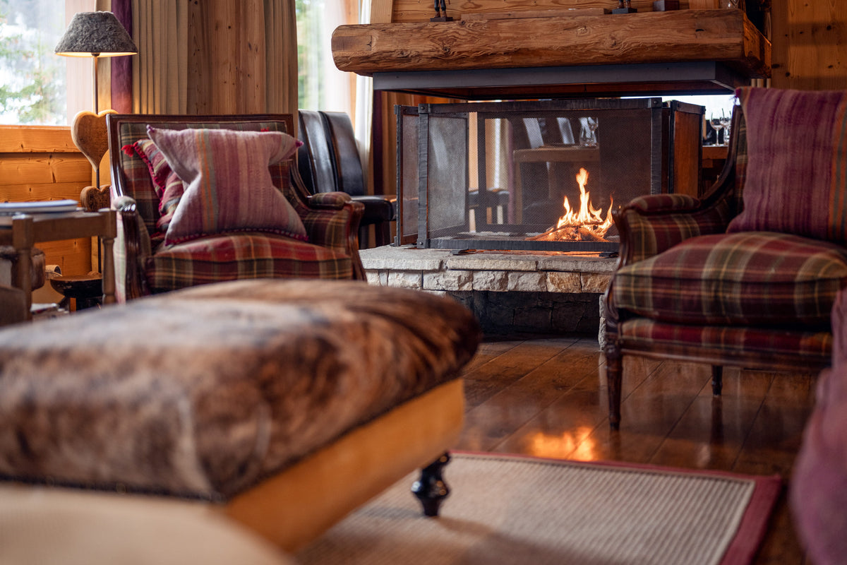 A cozy rustic living room scene with two plaid armchairs, a matching ottoman in the foreground, and a fireplace with a visible fire. The room has wood walls, a wood-burning fireplace, and warm ambient lighting.