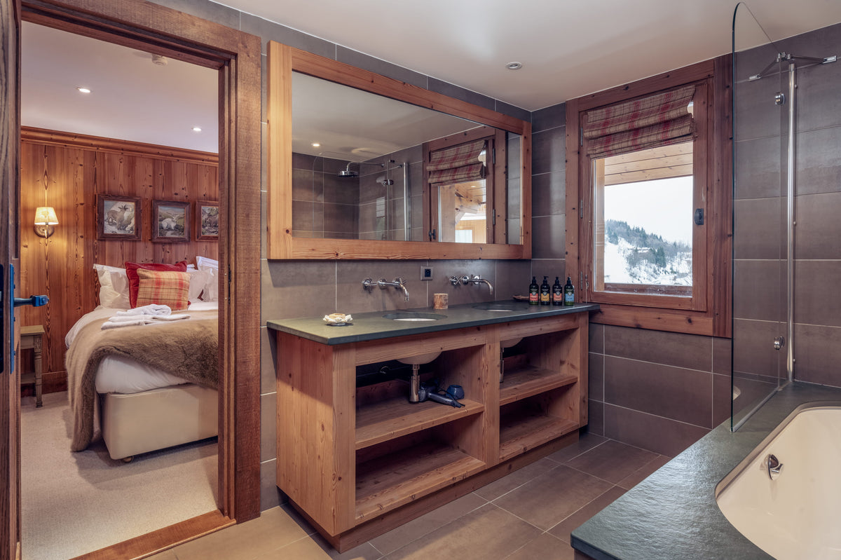 A luxury ski resort bathroom featuring a rustic wooden vanity with a dark stone countertop, a large wall mirror, and an open shower cabin with a glass door overlooking a snowy mountain landscape.
