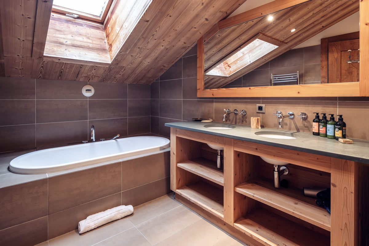 A luxury ski chalet bathroom featuring a large white freestanding bathtub set into a tiled wall, a double-sink vanity with open wooden shelving, and a large wood-framed mirror reflecting a skylight.