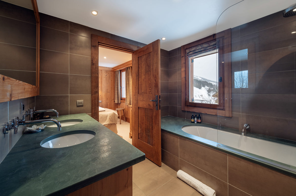 Luxury ski resort bathroom featuring a dark green stone double vanity with an oval sink, a white bathtub with a glass shower screen, dark brown tiled walls, and a wooden door opening to a bedroom with a snowy mountain view.
