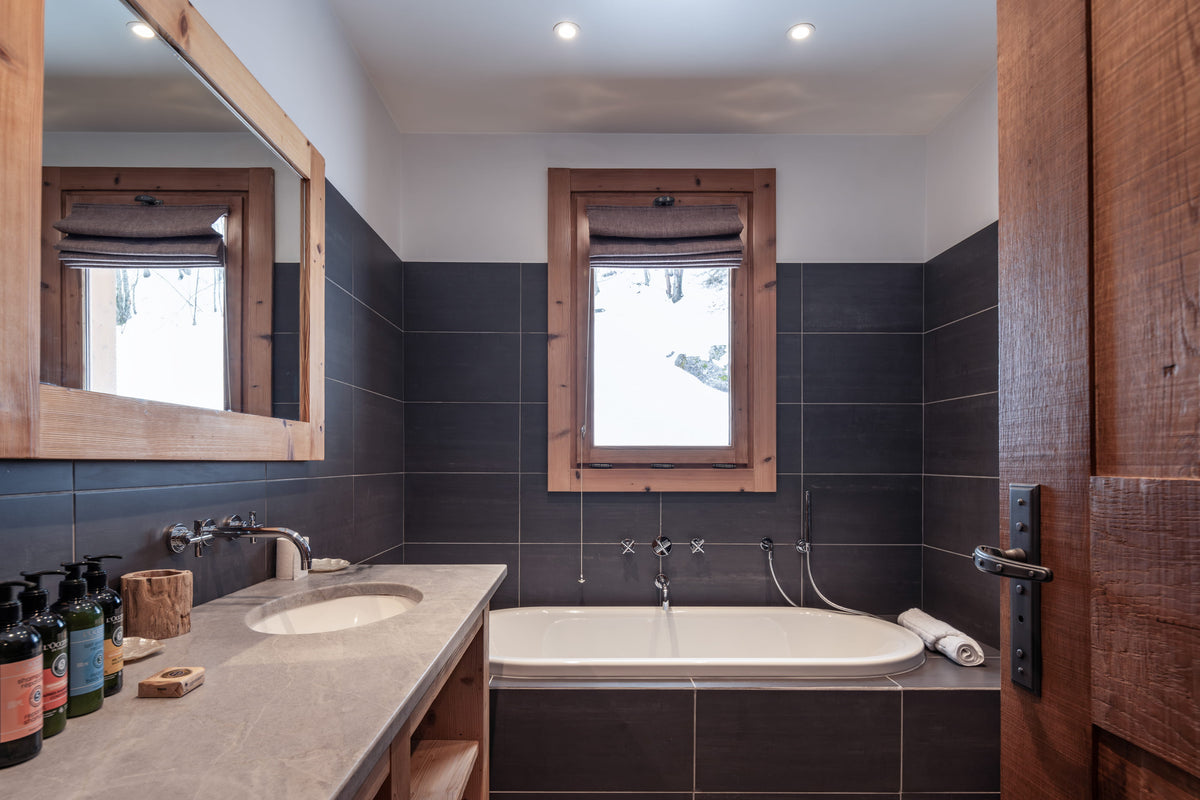 A modern bathroom interior with dark charcoal gray wall tiles, a white bathtub, a stone countertop with an integrated sink, a large wood-framed mirror, and a wooden door frame on the right.