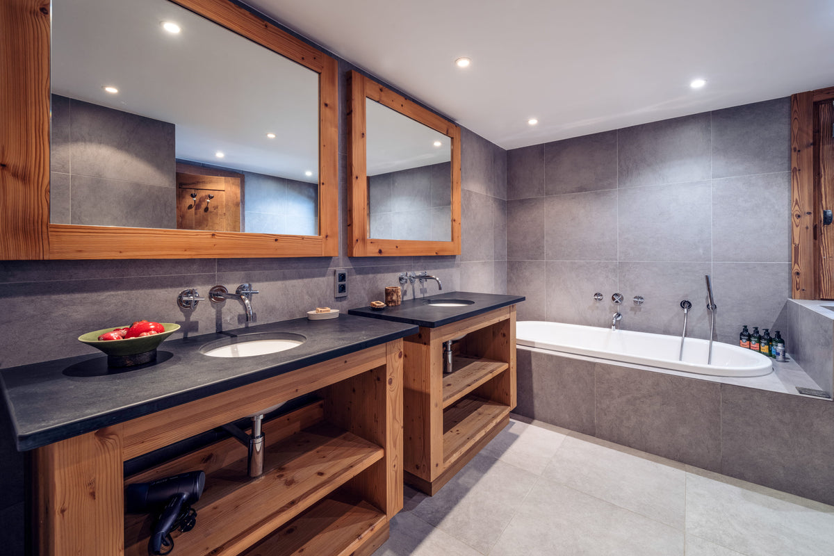 A modern bathroom interior with a large wooden-framed double vanity, two sinks, dark stone countertops, gray tiled walls, and an integrated white bathtub.