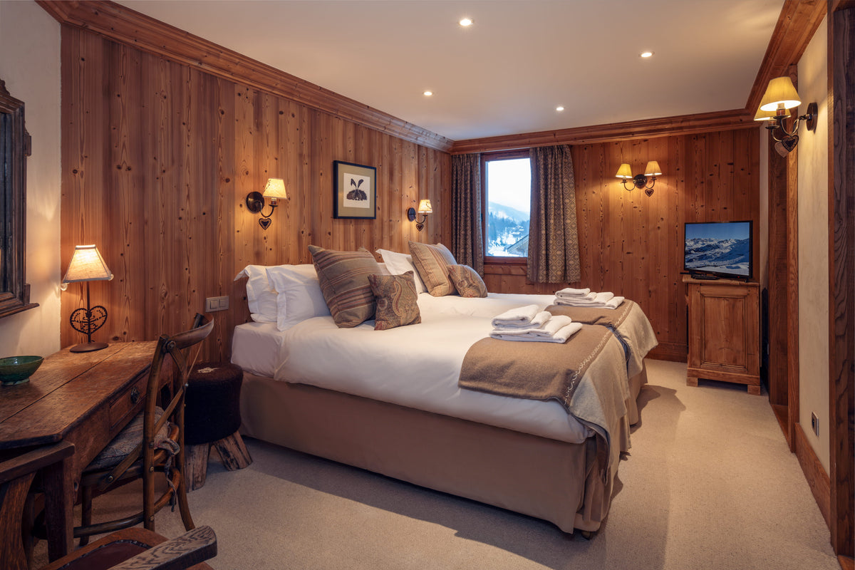 A cozy mountain-view bedroom with wood-paneled walls, a large bed dressed in white linens and tan bedding, bedside tables with lamps, and a window showing snowy scenery outside.