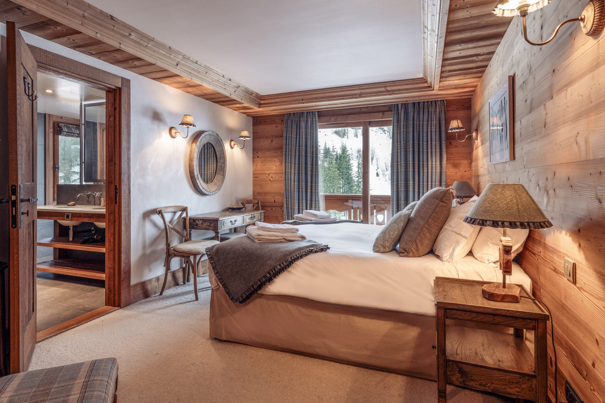 A cozy alpine-style bedroom with a large bed dressed in cream bedding, set against rustic wood-paneled walls. A wooden nightstand with a lamp sits beside the bed, and an open doorway on the left reveals a bathroom.