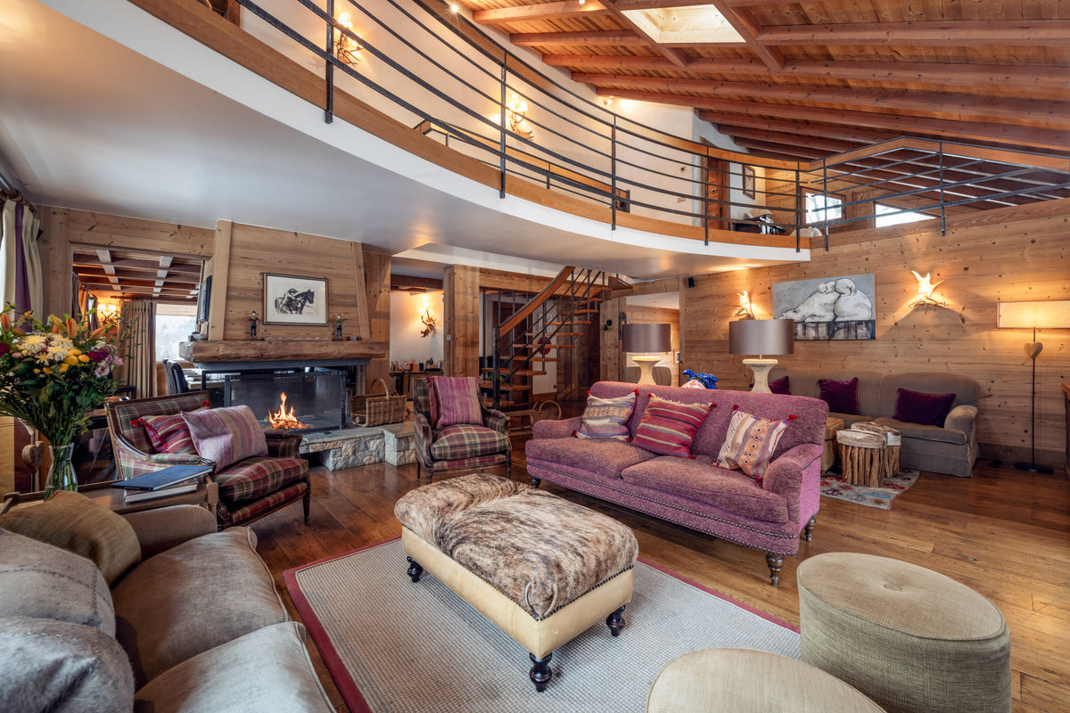 A rustic, cozy living room with a high wood ceiling and loft balcony, featuring a prominent purple sofa, a patterned ottoman, a fireplace, and warm ambient lighting.