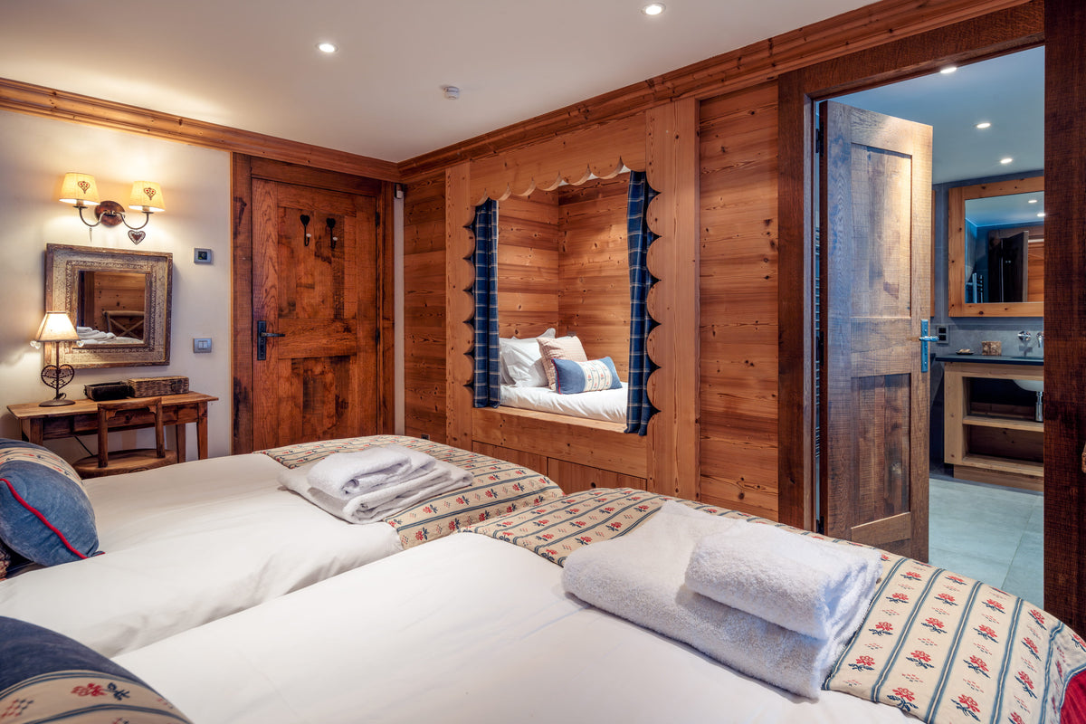 A cozy alpine-style bedroom with two single beds, white linens, patterned bed runners, wood-paneled walls, a built-in bench with a mirror, and an open doorway revealing a bathroom.