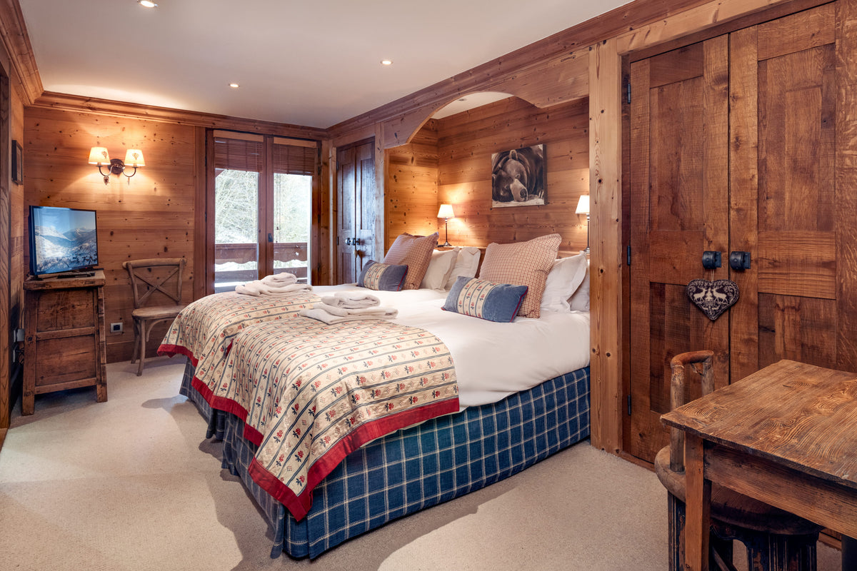 A rustic mountain-style bedroom with a large bed dressed in white linens and a patterned quilt, surrounded by warm wood-paneled walls, bedside lamps, a small TV, and a wooden desk.