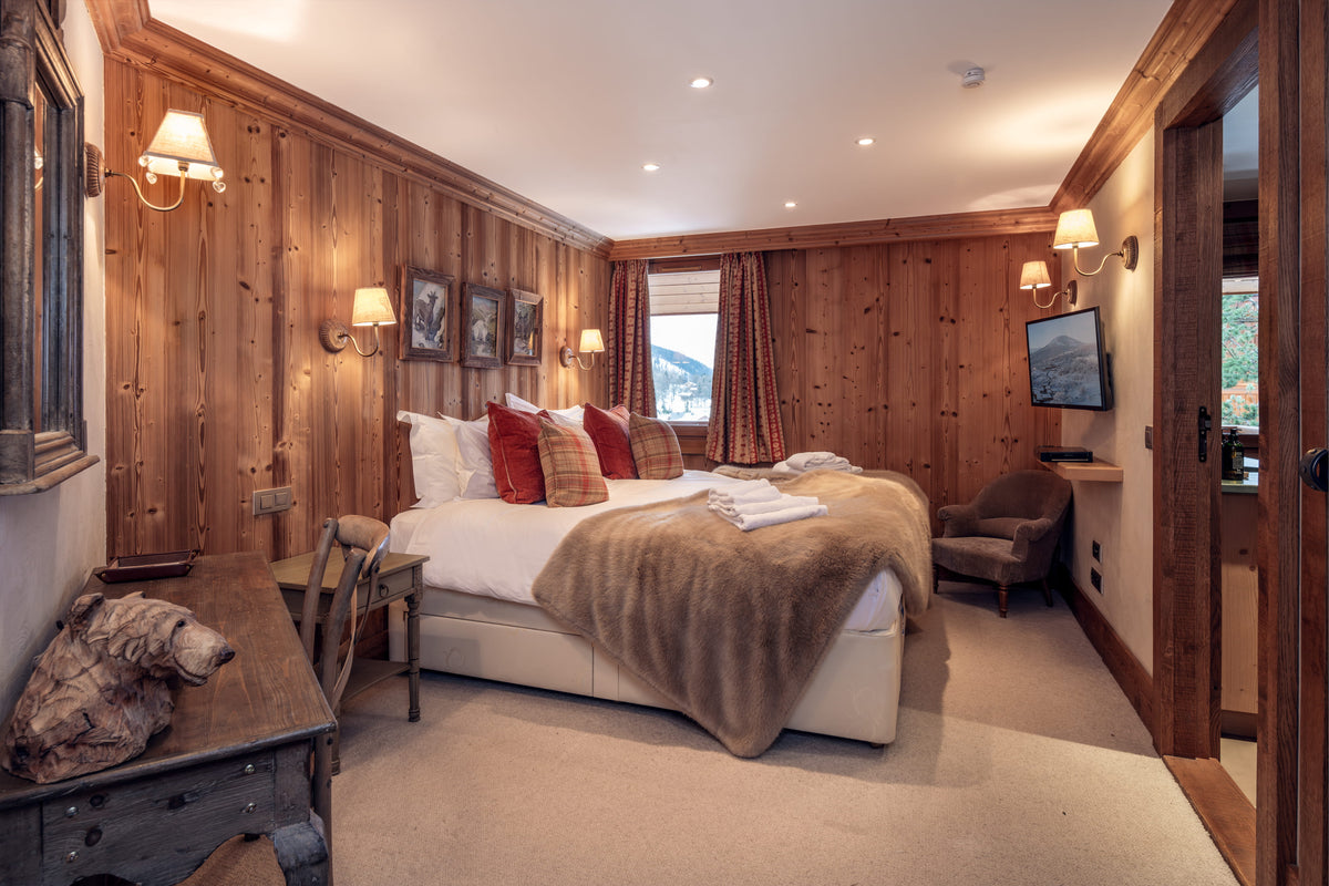 A cozy, rustic mountain chalet bedroom featuring warm wood-paneled walls, a large bed with white linens and a brown throw blanket, red accent pillows, a dark wood chest, and a wall-mounted television.