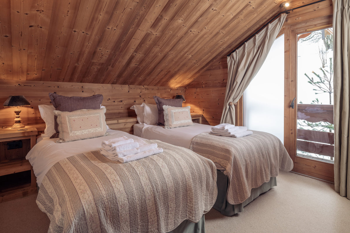 Two twin beds with beige and white striped bedding in a rustic wooden chalet bedroom, featuring a slanted wood ceiling, bedside tables with lamps, and a large window looking out onto a snowy outdoor scene.