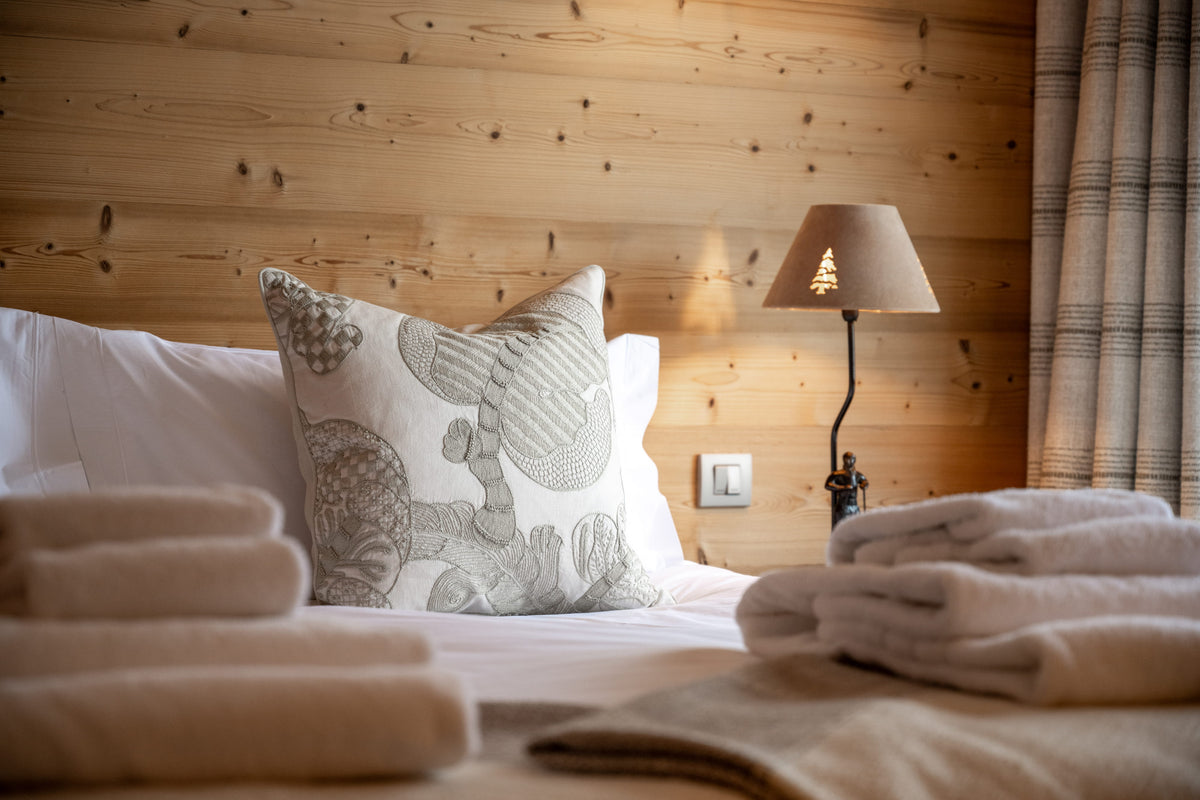 A cozy bedroom scene with a bed made of light wood, white bedding, a patterned throw pillow, folded towels, and a bedside lamp.