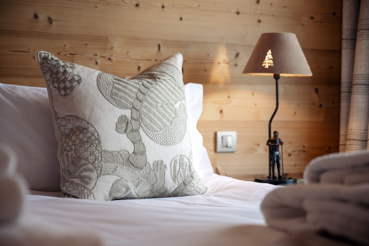 A decorative square throw pillow with a gray and white abstract pattern, resting on a white bed in a cozy bedroom with a wood-paneled wall, a lit lamp, and folded towels.