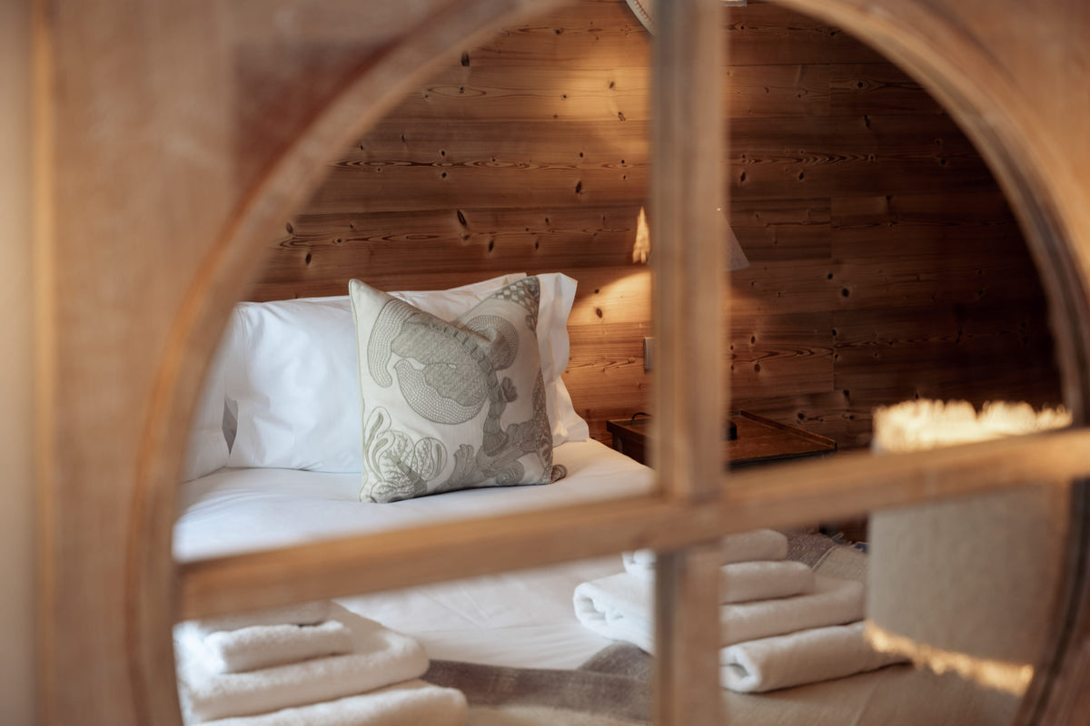 A cozy bedroom scene viewed through a rustic wooden arched frame, showing a neatly made bed with white linens and a patterned gray pillow against a warm wood-paneled wall.