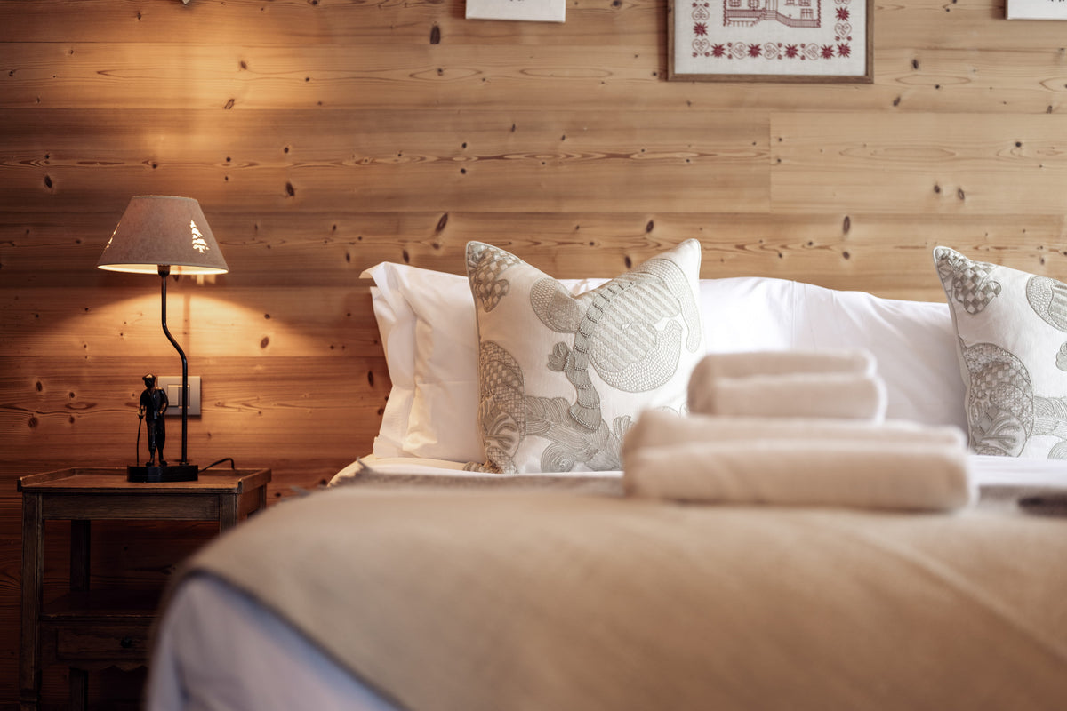A cozy bedroom scene with a neatly made bed in the foreground, white pillows and patterned accent pillows, folded towels, a bedside table, and a lit table lamp against a wood-paneled wall.