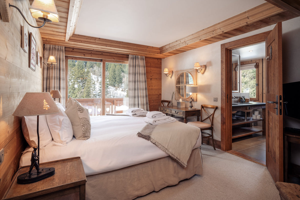 A cozy, rustic mountain-style bedroom featuring a large bed with white linens and beige bedding, wooden walls and ceiling beams, a bedside table with a lamp, and an open doorway revealing a bathroom.