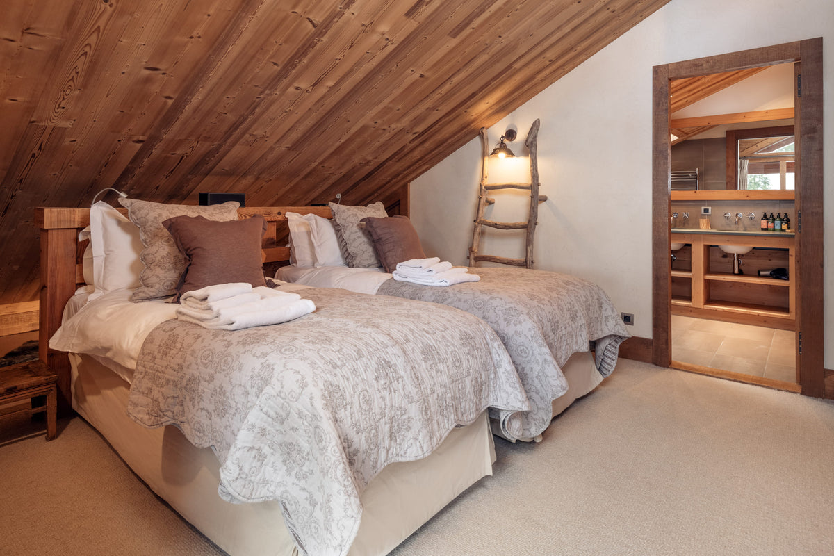 A rustic bedroom with two single beds under a sloped wooden ceiling, dressed in neutral beige and gray bedding. A wooden ladder-style wall lamp stands beside the beds, and an open doorway on the right reveals a wine bar area in the background.
