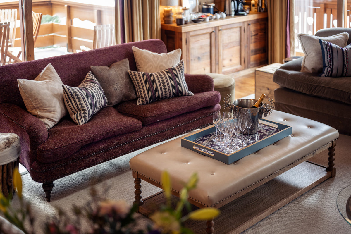 A cozy living room scene featuring a deep burgundy velvet sofa with patterned throw pillows, a cream tufted ottoman coffee table, and warm wood cabinetry in the background.