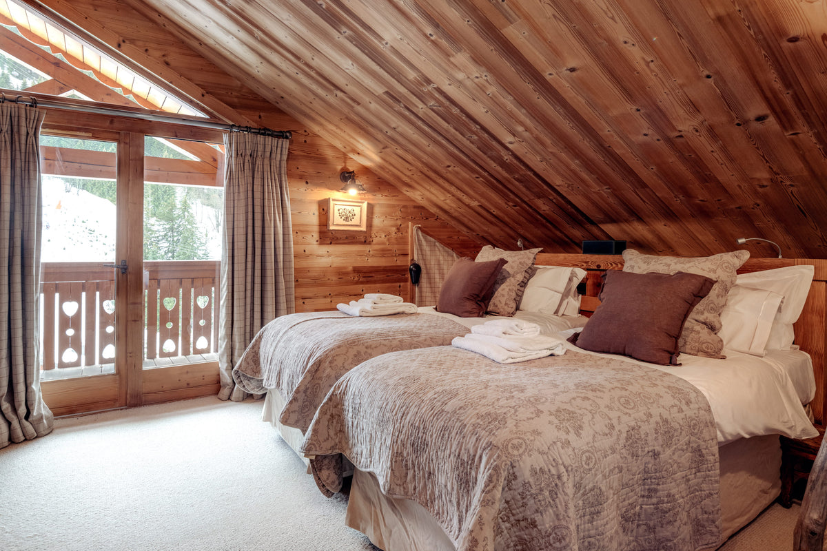 A cozy mountain-style bedroom with two neatly made beds, patterned beige and brown bedding, and dark brown pillows. The room features a rustic wooden slat ceiling and walls, with a glass door on the left leading to a balcony.