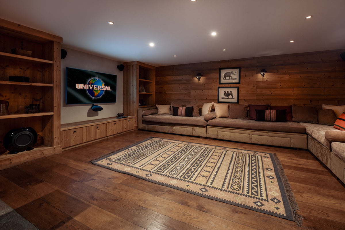 A cozy home theater setup with a large sectional sofa, built-in wooden shelves, a TV displaying the Universal logo, and a patterned area rug on a hardwood floor.