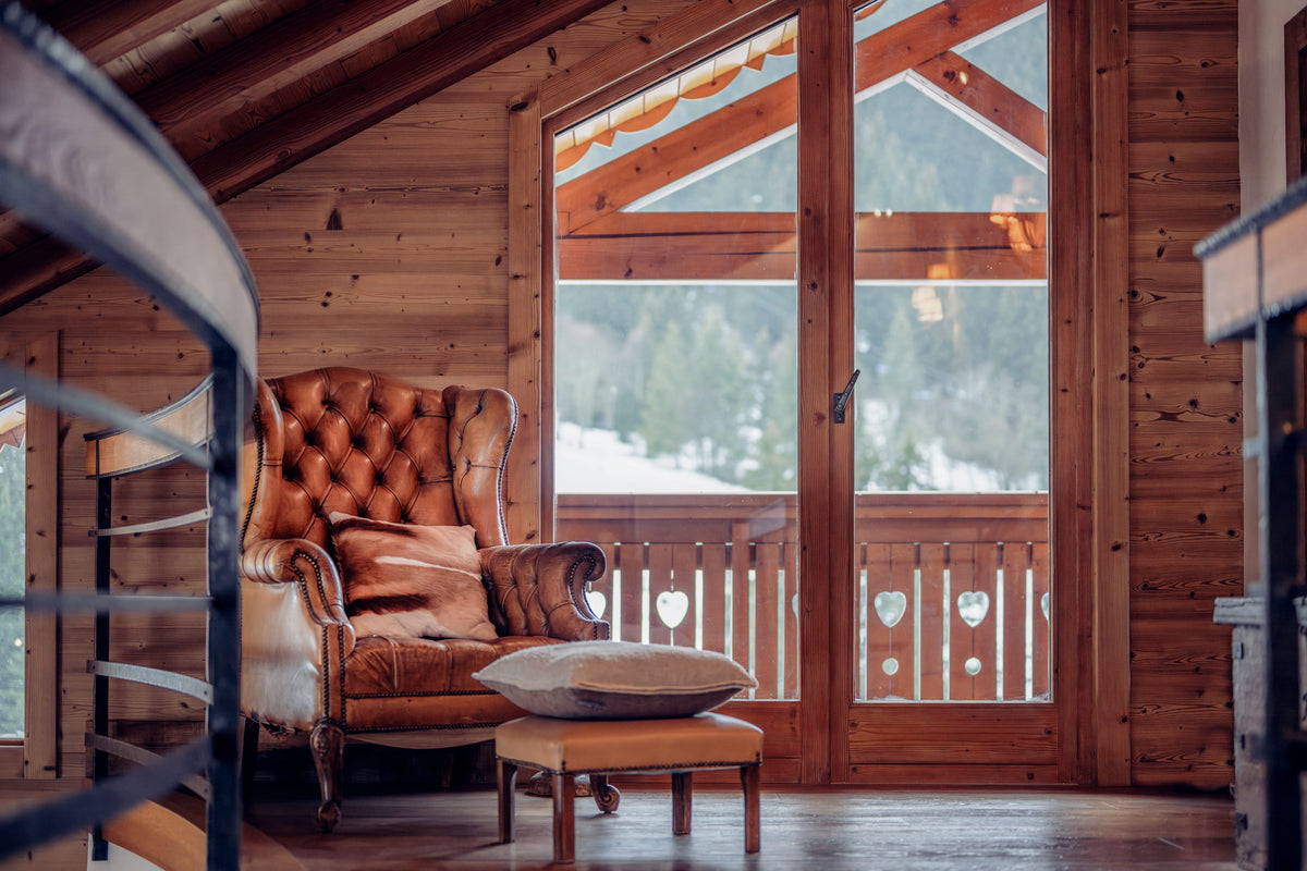 A warm brown tufted leather armchair with matching cushions sits in a rustic wooden chalet interior, with large glass doors showing a snowy outdoor view and a small wooden ottoman in front.