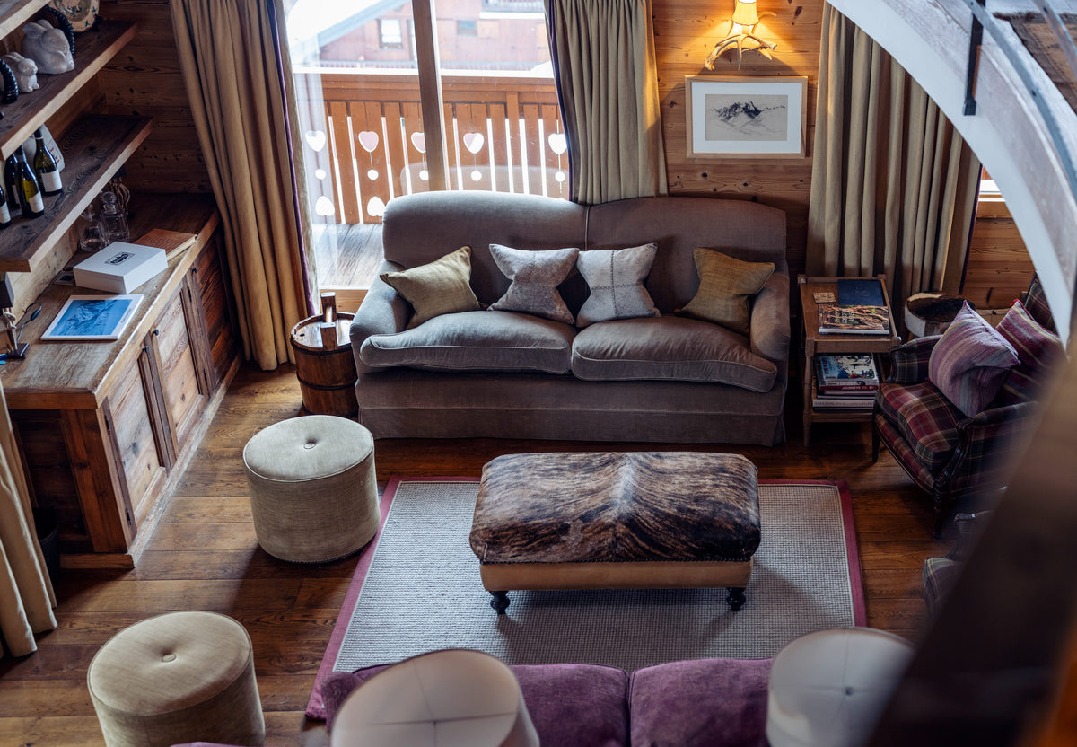 A cozy, rustic chalet-style living room featuring a gray fabric sofa with decorative pillows, a patterned ottoman, wooden shelves, and a large window looking out to a wooden balcony.