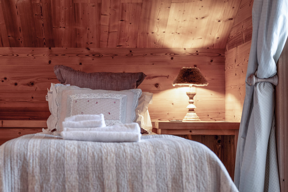 A cozy bedroom scene in a rustic wood cabin with a neatly made bed, white folded towels, a lit table lamp, and light blue curtains tied back on the right.