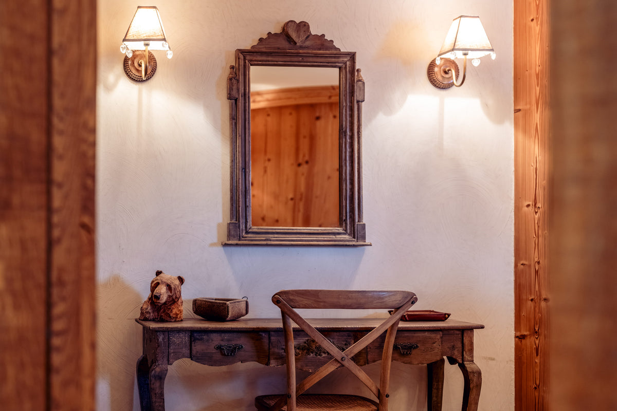 A rustic interior scene with a dark wood console table, matching chair, wall mirror, and two lit sconces against a warm off-white wall.