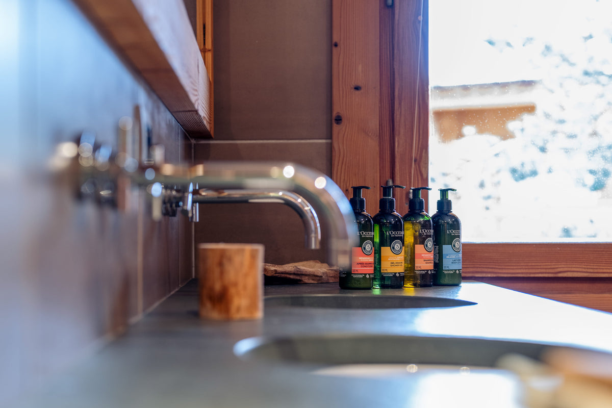 Four dark green pump bottles of olive oil with black caps lined up on a dark countertop next to a window in a rustic kitchen setting.