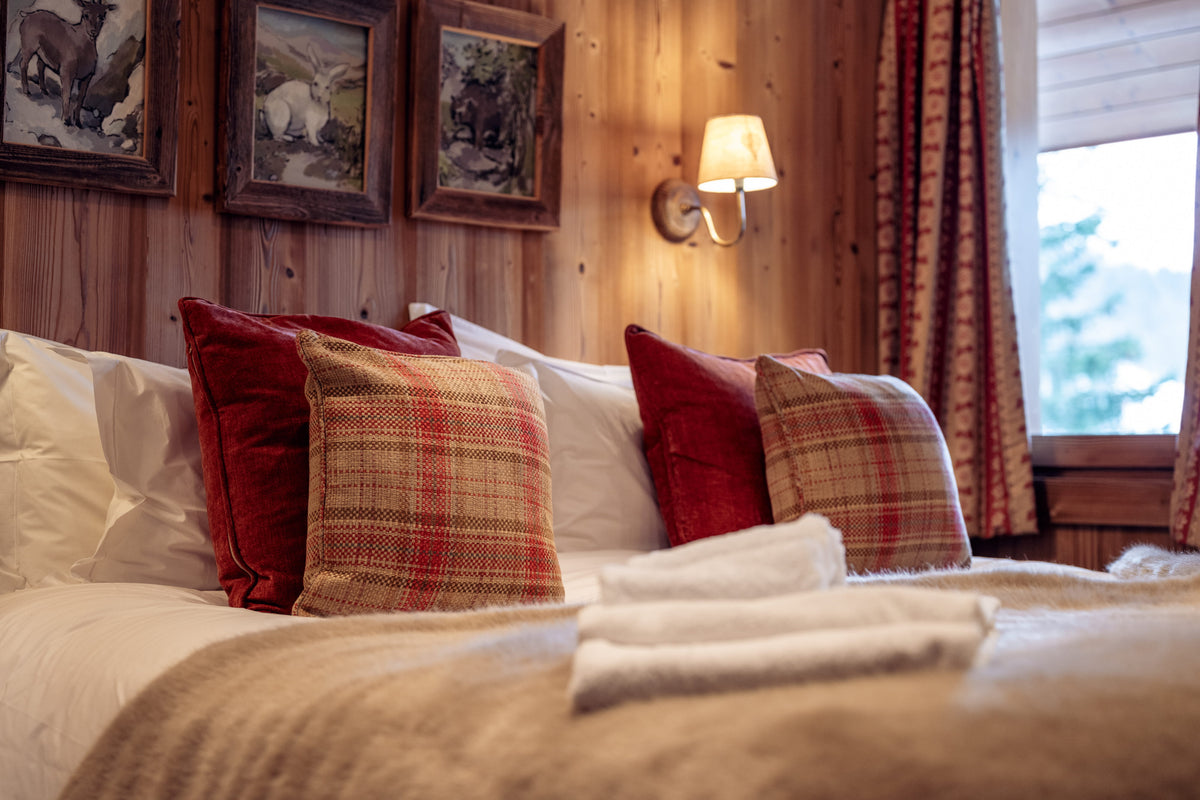 A cozy bedroom scene with a neatly made bed in a wood-paneled room, featuring red and plaid accent pillows, white bedding, a lit wall sconce, and framed artwork above the headboard.