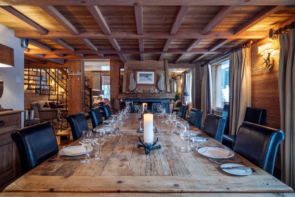 A rustic wooden dining table set with white plates, wine glasses, and a central white pillar candle, surrounded by black leather chairs in a cozy, wood-paneled chalet interior.