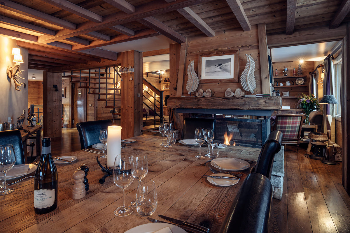 A rustic alpine chalet dining room with a wooden dining table set for a meal, a wine bottle, and candles, with a fireplace and wooden ceiling beams in the background.