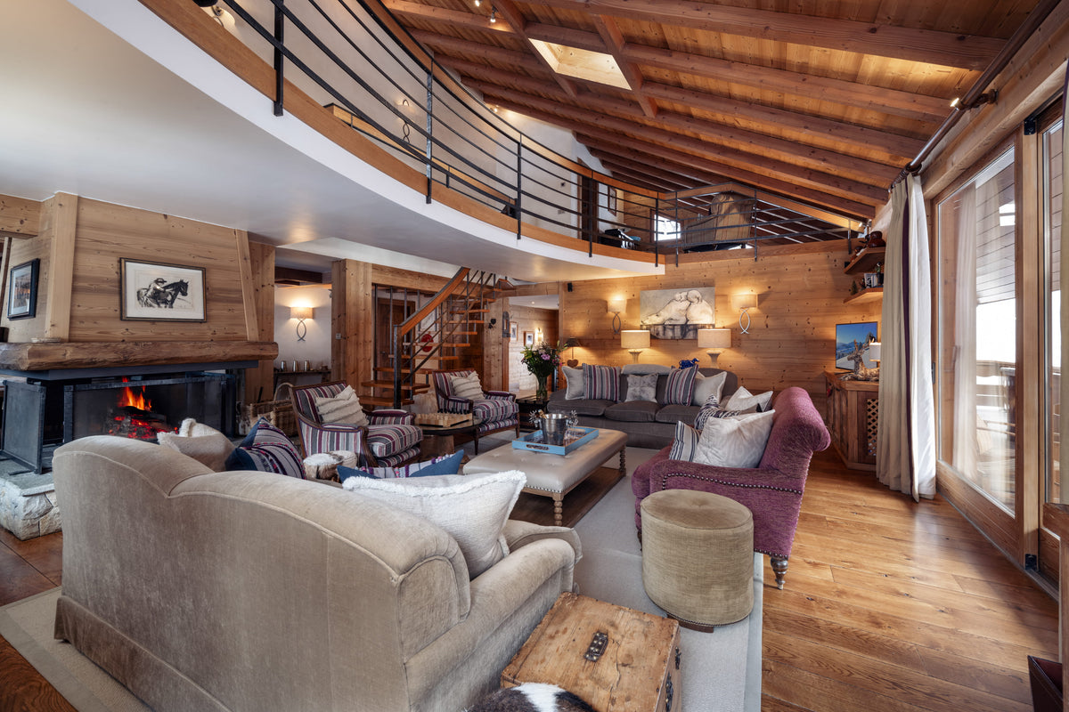 A luxurious mountain chalet living room with a high wood ceiling, featuring a beige velvet sofa in the foreground, a purple armchair, a fireplace with a wood-burning stove, and a loft balcony with a black metal railing.