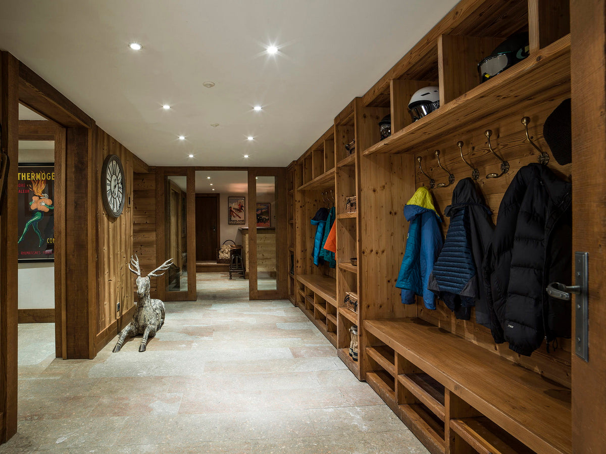 A long wooden ski lodge entryway with built-in shelving, coat hooks, bench seating, and several winter jackets hanging on the right. A deer statue stands near the left wall, and the space is lit by recessed ceiling lights.