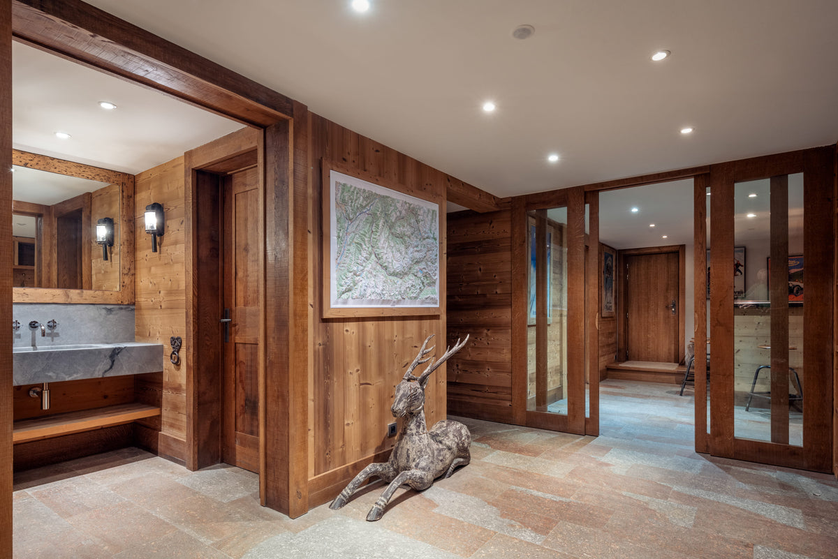 A rustic lodge-style interior hallway with wood-paneled walls, recessed ceiling lights, stone tile flooring, a framed landscape print, and a deer sculpture near the center.