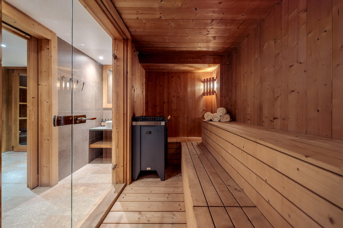A wooden sauna interior with tiered seating, wood-paneled walls and ceiling, a glass door on the left, and a dark gray freestanding appliance near the center.