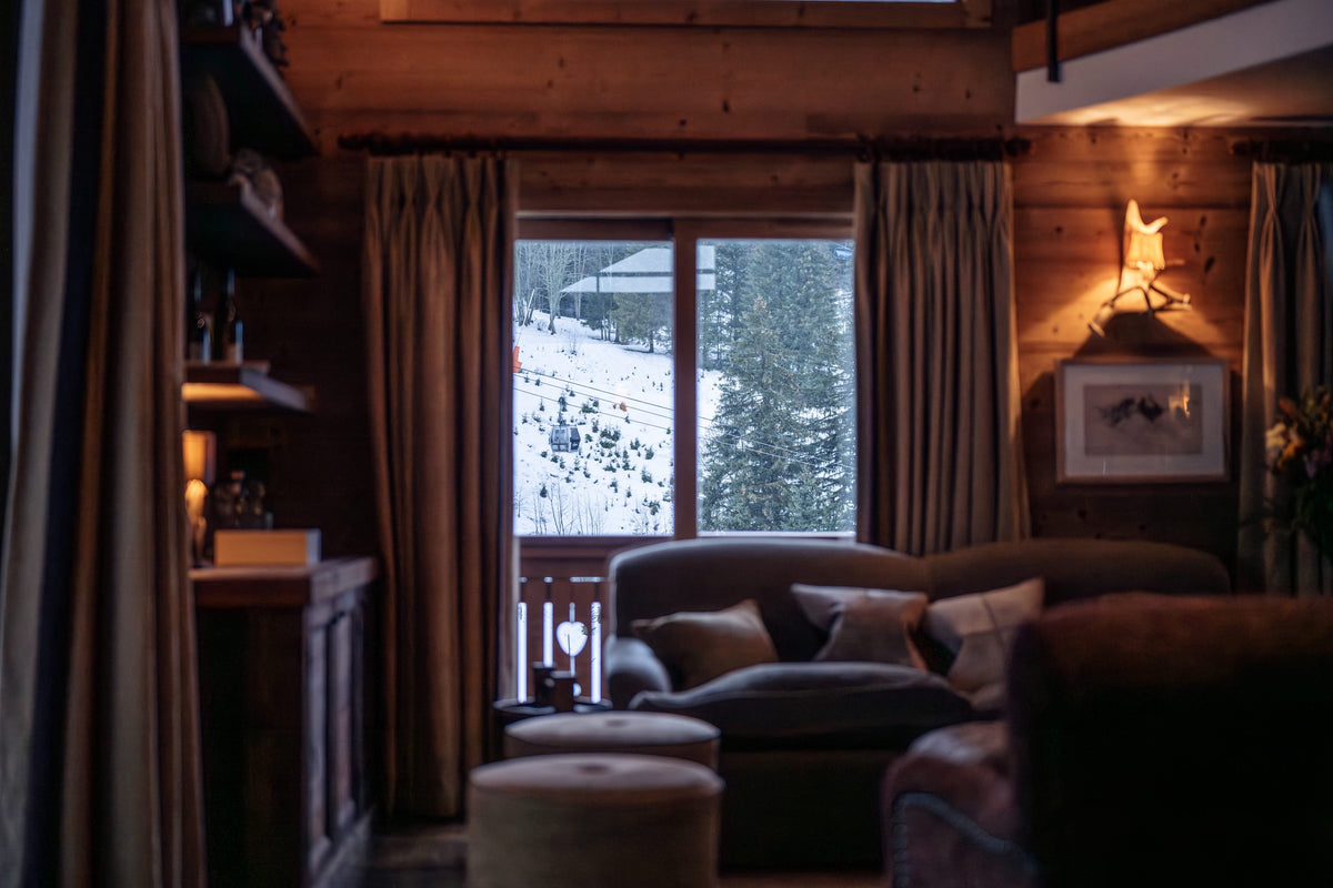 A cozy, warmly lit mountain chalet living room with wood-paneled walls, a dark gray sofa with pillows, and a large window looking out onto a snowy mountain landscape.