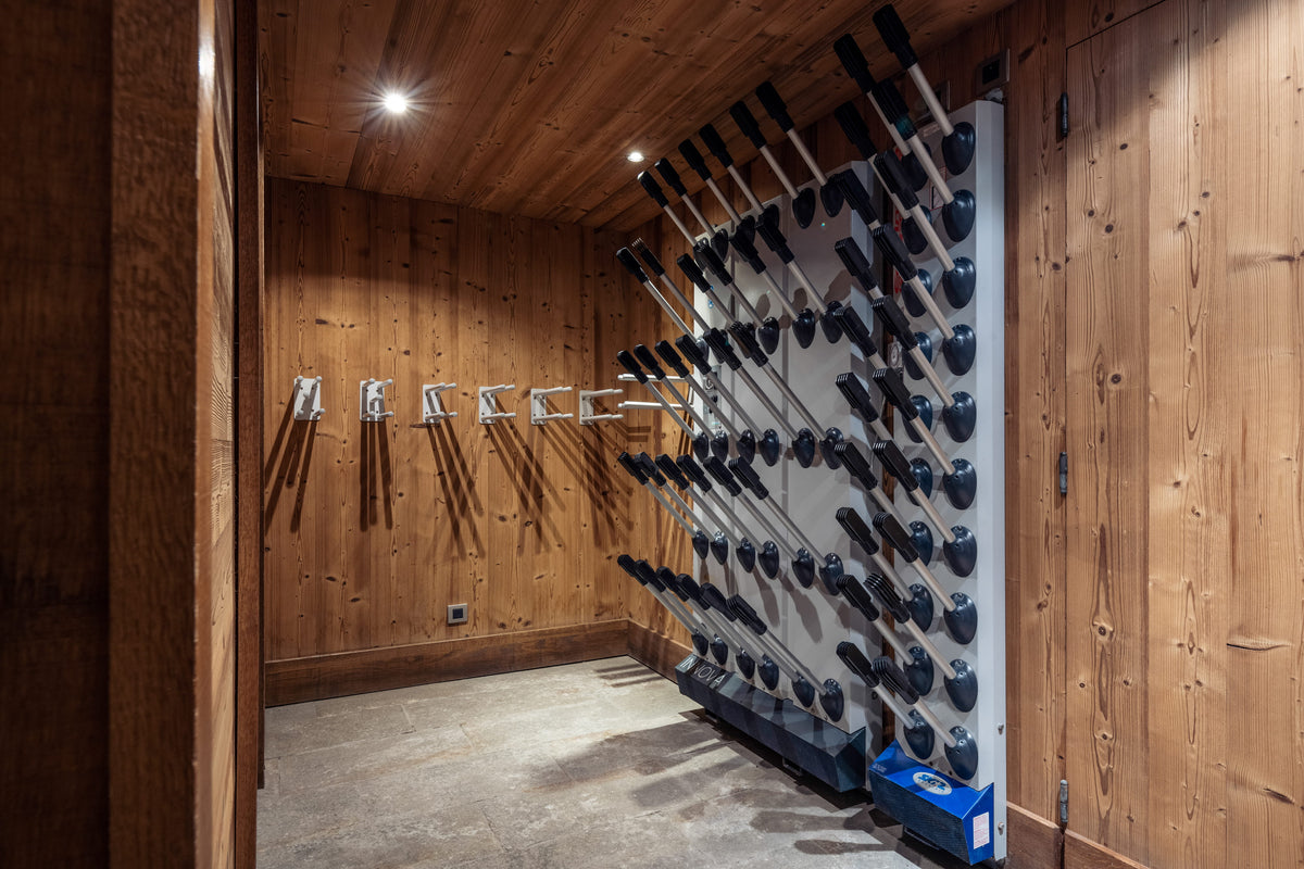 A wall-mounted rack filled with ski poles and ski boots in a wood-paneled room with a concrete floor.