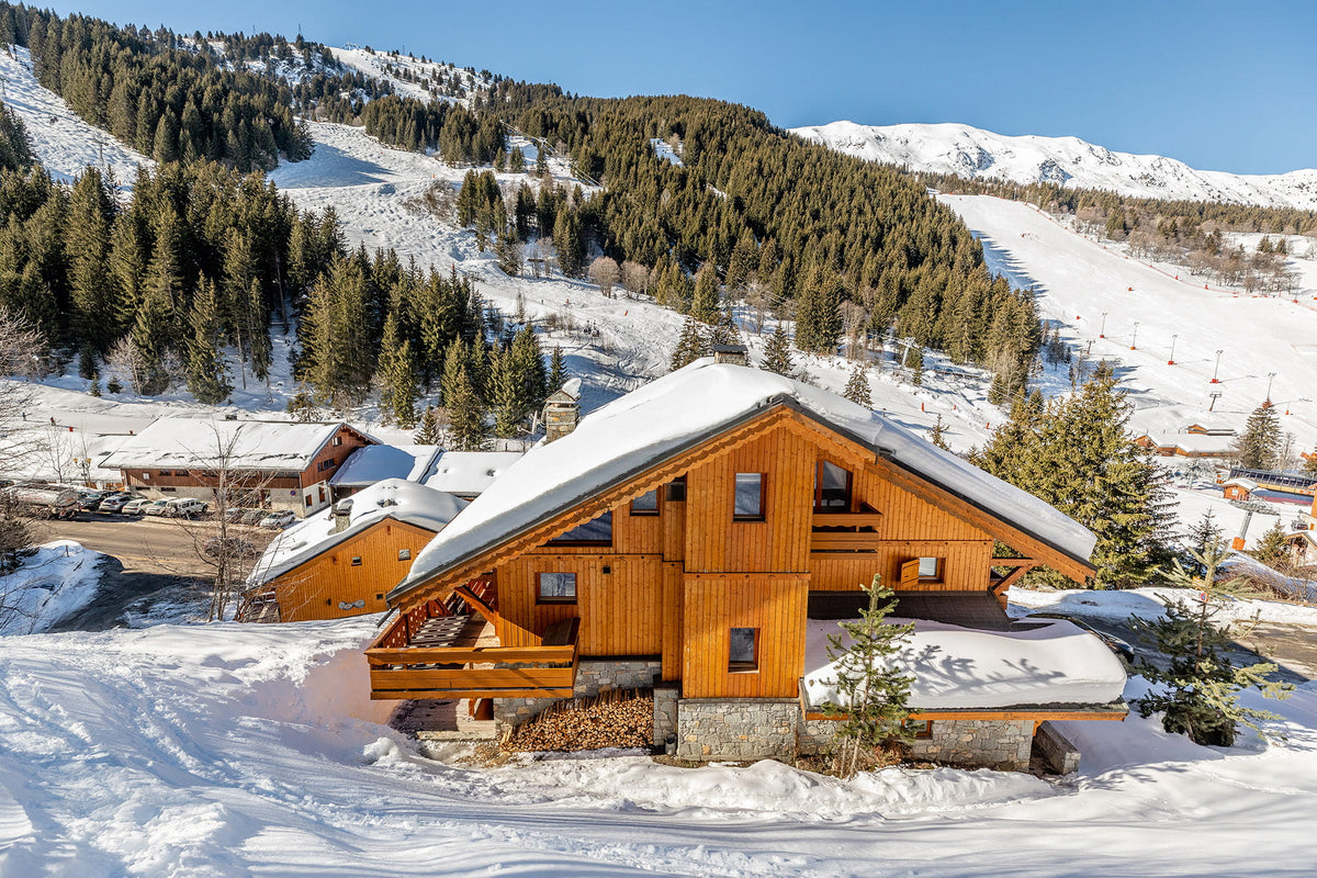 A wooden chalet with snow-covered roof and porch stands in a snowy mountain village, with evergreen trees and ski slopes in the background under a clear blue sky.