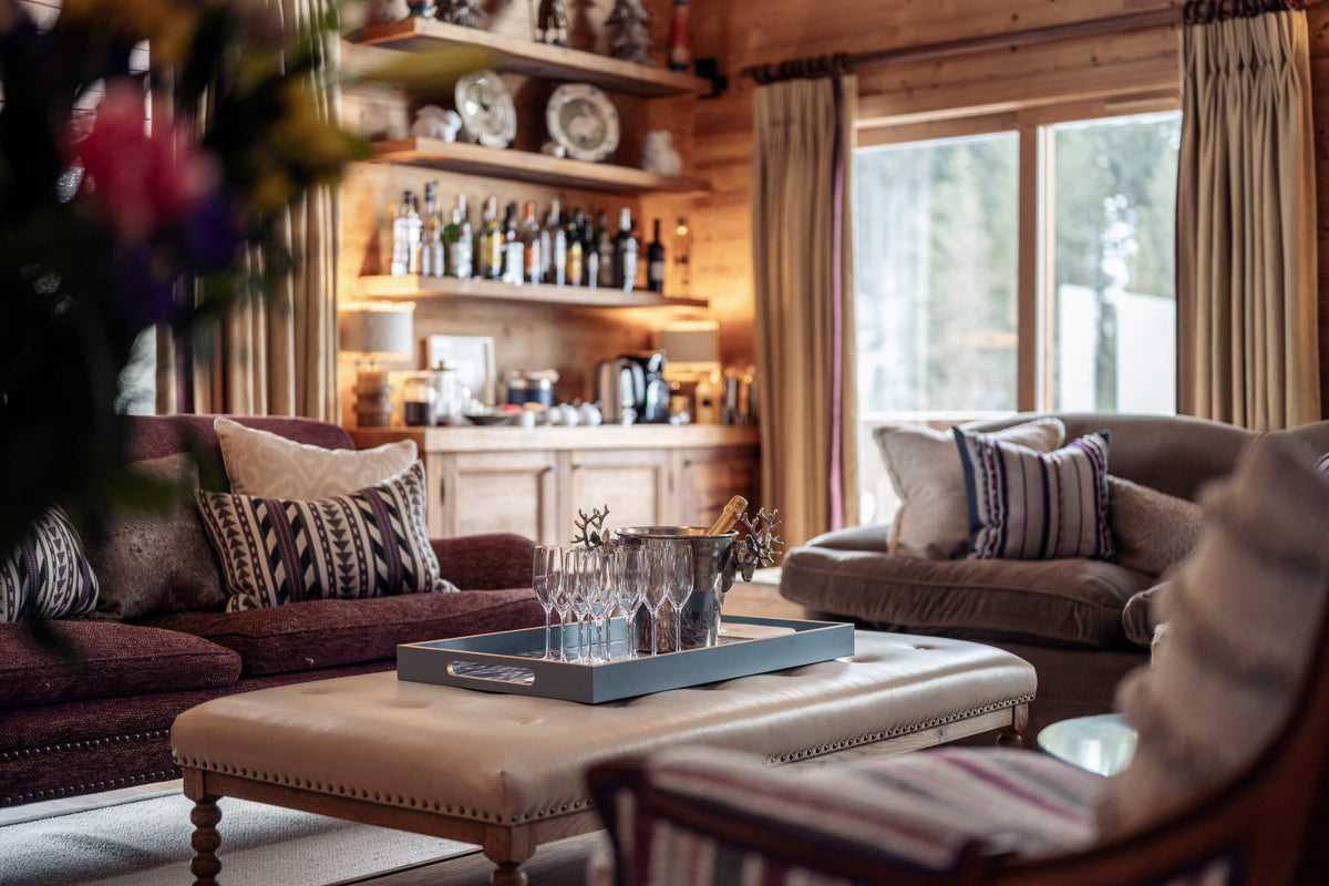 A cozy, rustic living room with warm wood-paneled walls, a built-in bar shelf with bottles, and plush sofas with patterned pillows. A cream-colored tufted coffee table with a blue tray sits in the foreground, and a large window with beige curtains lets in natural light.