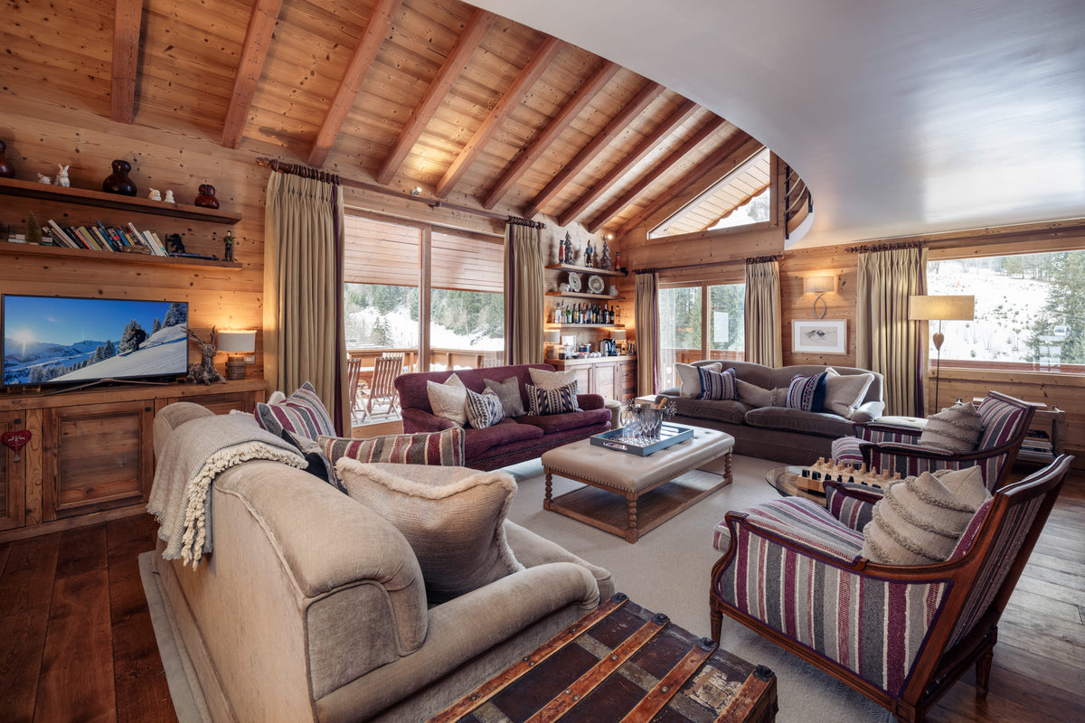 A luxurious ski chalet living room with a high arched wooden ceiling, featuring multiple plush sofas, striped armchairs, a wooden coffee table, and a large TV mounted on the wood-paneled wall.