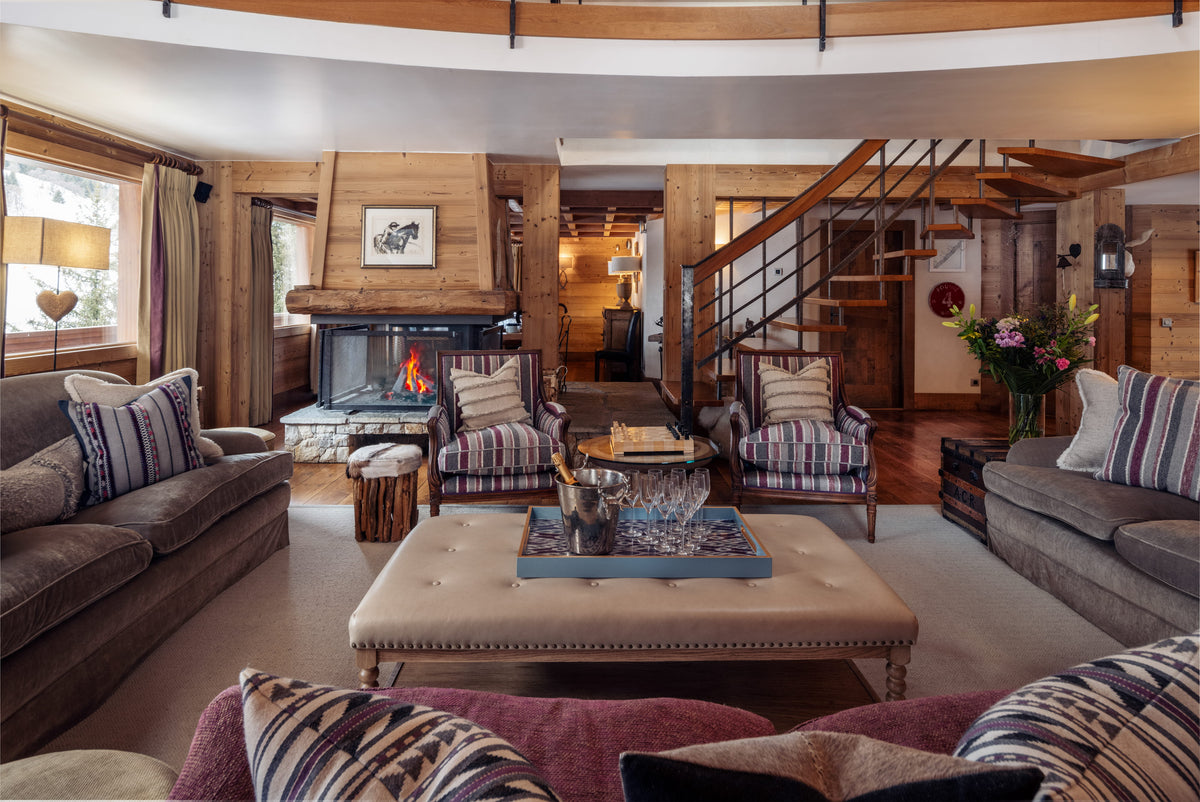 A cozy, rustic mountain-style living room with a large tufted beige coffee table in the foreground, striped accent chairs, brown sofas, a stone fireplace, and a wooden staircase in the background.