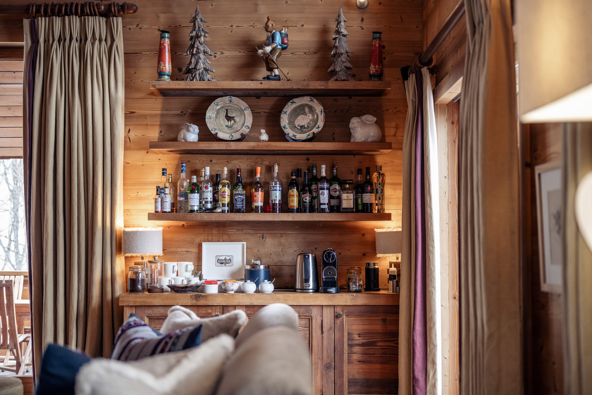 A rustic wooden bar cabinet with open shelves stocked with liquor bottles, decorative figurines, and two round wall clocks, set in a warm wood-paneled room with curtains and a sofa in the foreground.