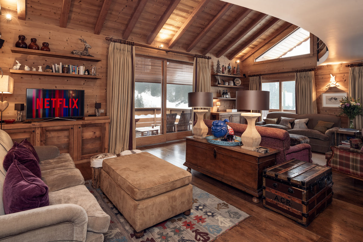 A cozy, rustic mountain chalet living room with wood-paneled walls and ceiling, featuring a beige sofa with purple pillows, a large tan ottoman, a wooden coffee table, and a television displaying the Netflix logo.