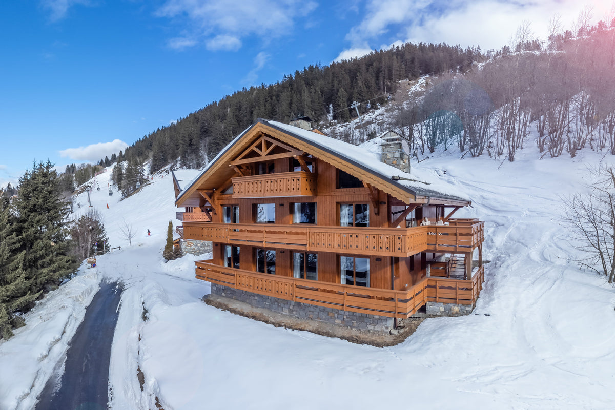 A wooden chalet-style mountain cabin with snow-covered roofs and balconies, situated on a snowy ski slope with evergreen trees and a blue sky in the background.
