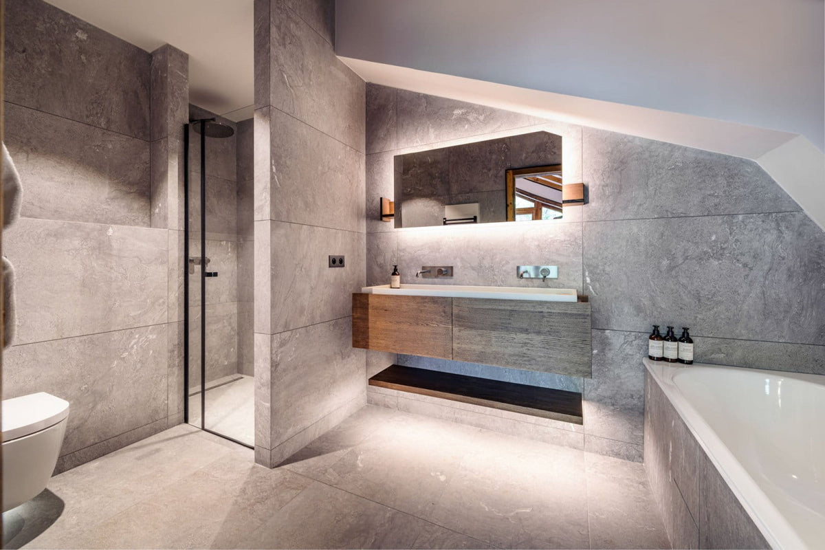 Modern bathroom interior with gray stone tile walls and floor, a floating wooden vanity with a white sink, a large mirror, a glass shower enclosure, a toilet on the left, and part of a bathtub on the right.